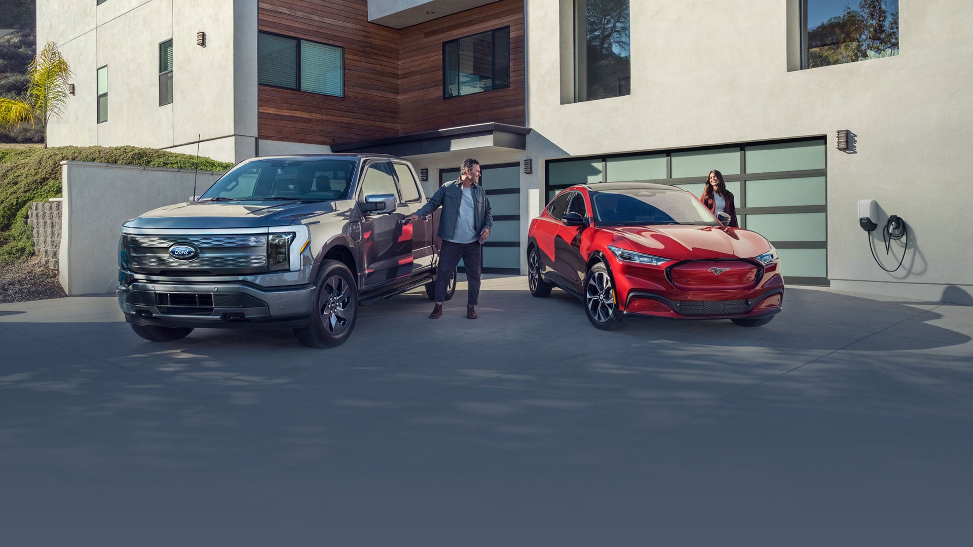 Grey F-150 Lightning and red Mustang Mach-E at a modern house with a man, woman, and car charger.