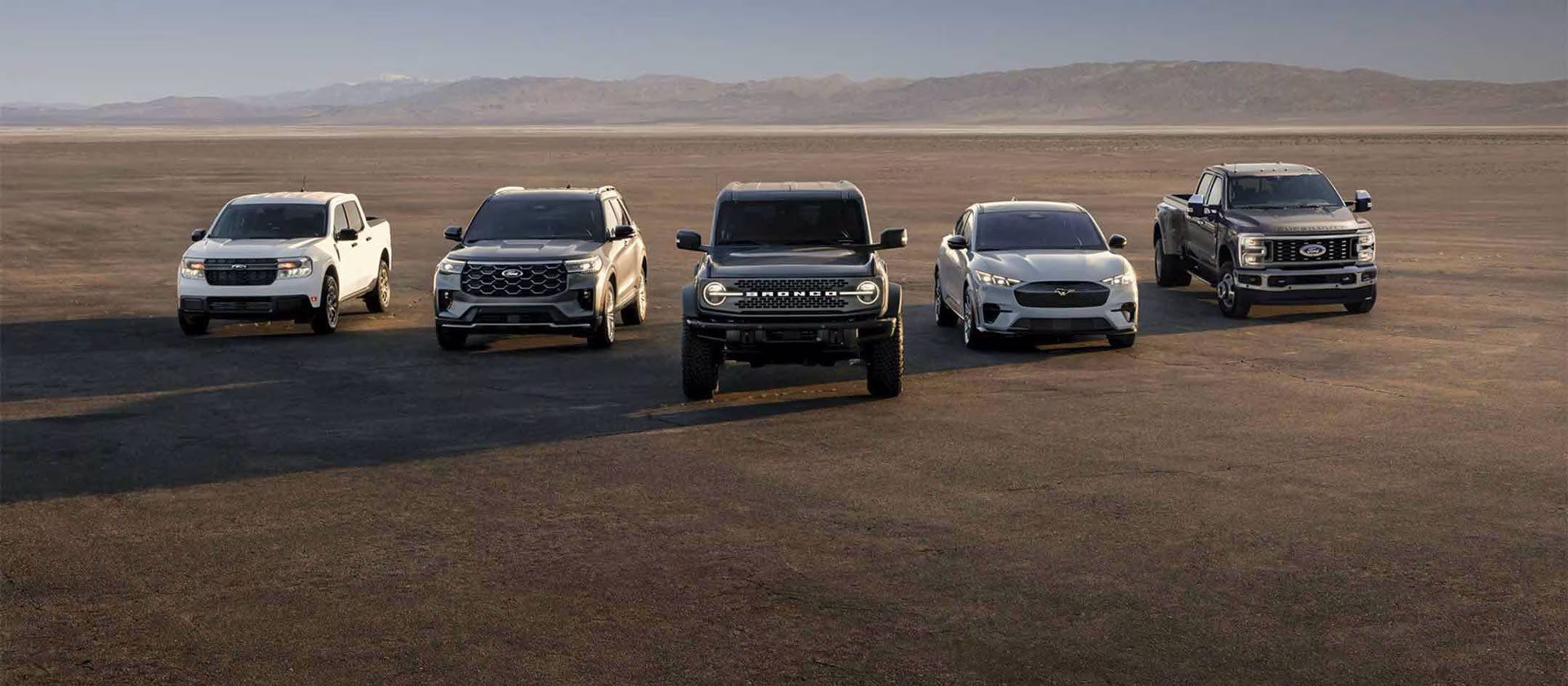 Five Ford vehicles—Maverick, Explorer, Bronco, Mach-E, Super Duty—parked in a line on a desolate plain with mountains.