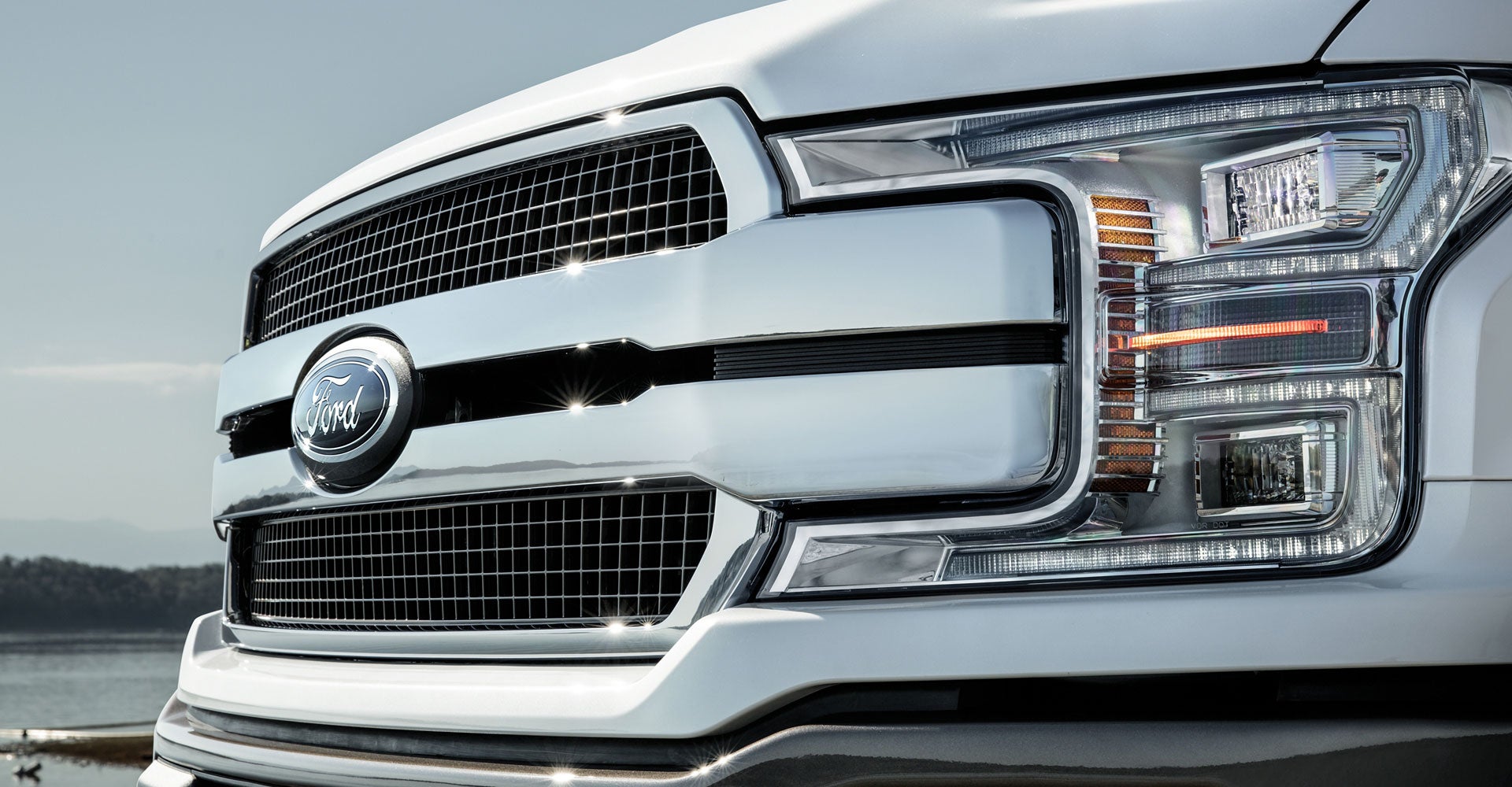 Close-up of a white Ford truck's front with a large chrome grille, blue Ford logo, and complex LED headlights.