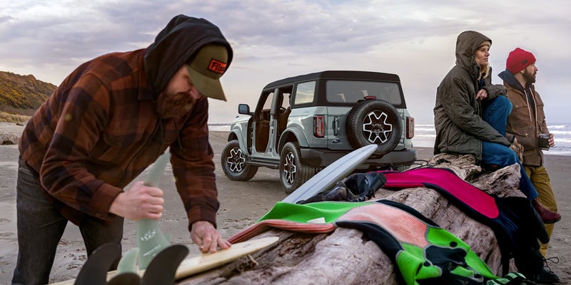 Man preparing surfboard on a beach with a gray Ford Bronco, doors off, and two people sitting on driftwood watching the ocean.