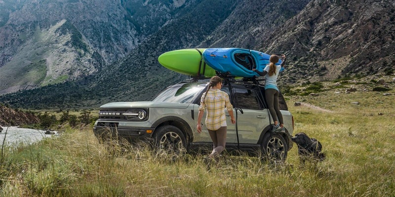Two people load colorful kayaks onto a grey Ford Bronco Sport SUV in a grassy mountain landscape.