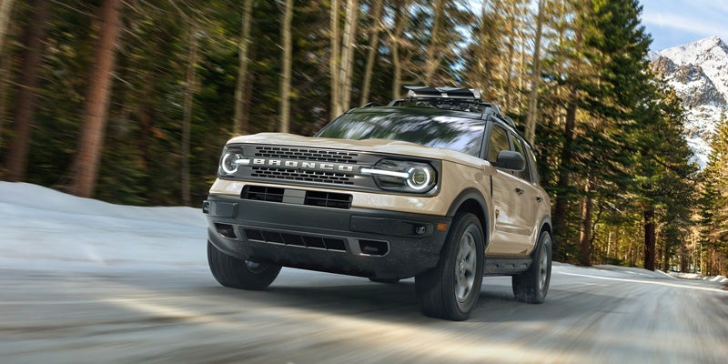 Tan Ford Bronco Sport SUV with roof rack driving on a snowy road through a forest with mountains.