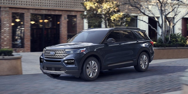 Dark gray Ford Explorer SUV drives on a paved street past brick buildings and trees.