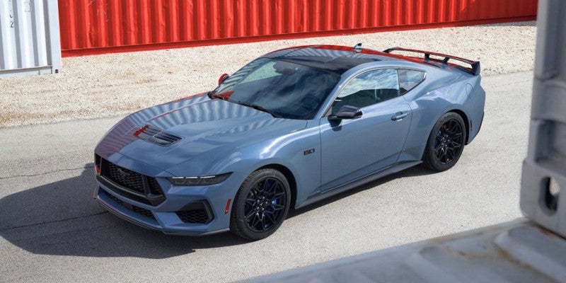 A blue-grey 2024 Ford Mustang sports car with black wheels and spoiler, parked on asphalt near red shipping containers.