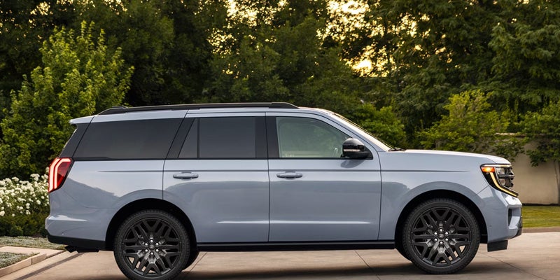 Side profile of a light gray Ford Expedition SUV parked on a paved driveway with trees