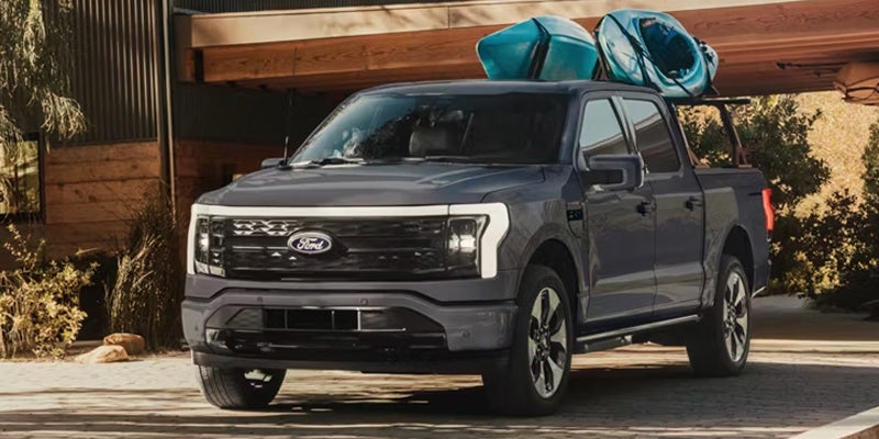 Dark gray Ford F-150 Lightning pickup truck with two blue kayaks on the roof rack, parked in front of a modern house.