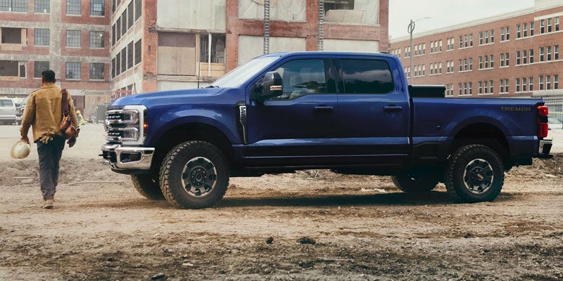 Blue Ford F-150 pickup truck towing a large black travel trailer while driving on a tree-lined road.