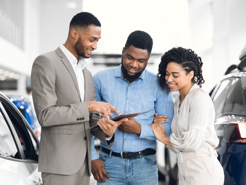 Car salesman points to a document for a smiling couple at a car dealership.