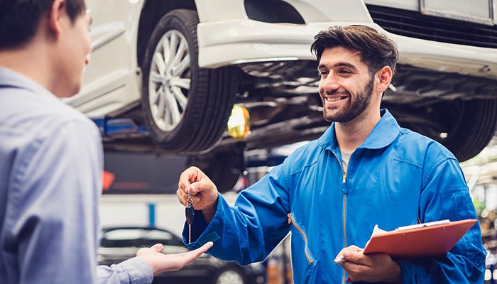 Smiling mechanic in blue hands car keys to a customer in a garage with a car on a lift.